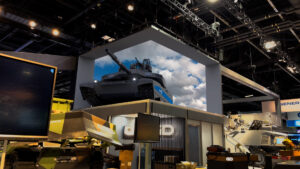 A large armored military tank showcased on a display platform at a trade show, with General Dynamics branding visible. The tank is set against a backdrop of a clear blue sky with clouds.