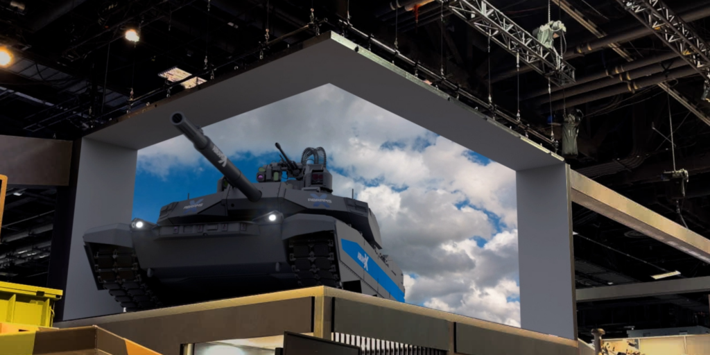 A large armored military tank showcased on a display platform at a trade show, with General Dynamics branding visible. The tank is set against a backdrop of a clear blue sky with clouds.