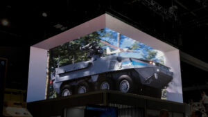 An anamorphic screen display showcasing a military vehicle firing in a dynamic action scene at a defense exhibition.