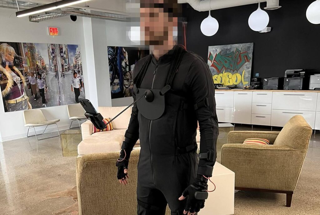 A person wearing a full-body motion capture suit with sensors on their arms, legs, and gloves, standing in a modern office space with graffiti-style artwork and lounge furniture in the background.