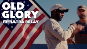 A Team RWB runner, wearing a cap and sunglasses, hands off a baton during the Old Glory Ultra Relay while carrying an American flag. The text "OLD GLORY ULTRA RELAY" is prominently displayed over the flag, symbolizing the endurance and patriotism of this historic event.