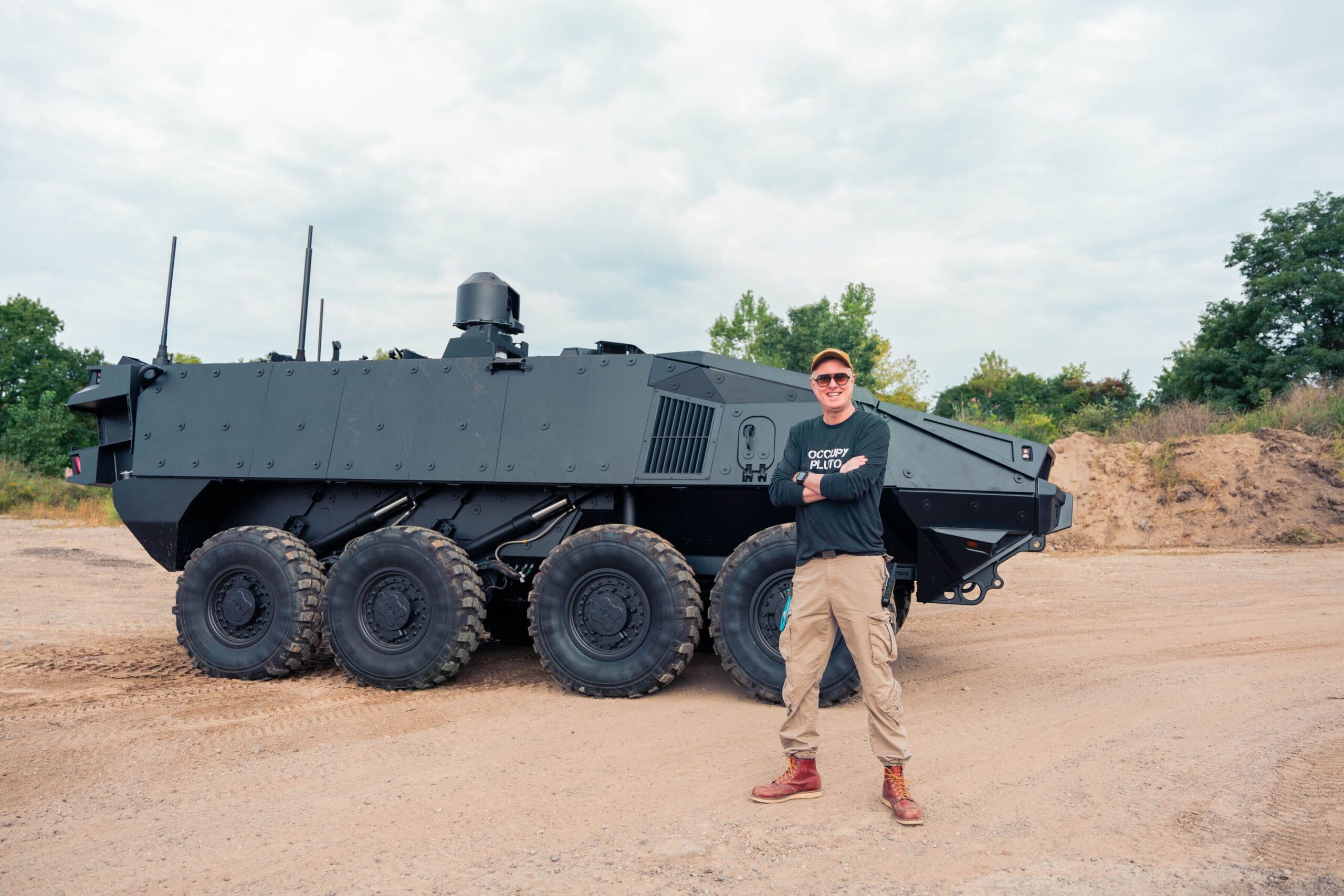 Man standing in front of an armored military vehicle on a dirt testing ground.